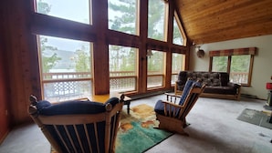 A spacious room with large windows, wooden furniture, and a view of trees and a deck outside.