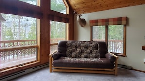 A wooden cabin interior with a sofa, large windows, a deer head mounted on the wall, and a view of trees.