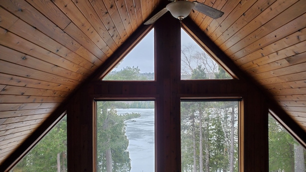 A room with a sloped wooden ceiling and a ceiling fan. There are four windows providing views of a forest and a river.