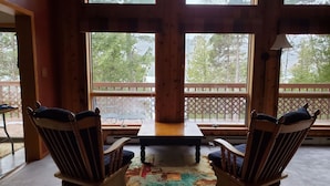 A wooden interior with a large window overlooking a deck, two wooden chairs, and a table.