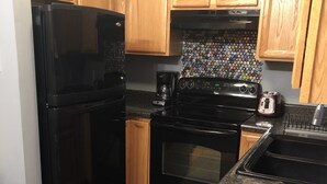A kitchen with wooden cabinets, a black refrigerator, a coffee maker, a toaster, and a backsplash made of bottle caps. - Silver Lake Cabin (Traverse City)