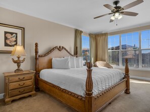 A bedroom with a wooden four-poster bed, a nightstand with a lamp, a framed picture on the wall, and a view of the outside through large windows. - Unique Corner Condo | Free Beach Service | Large Balcony | Designated Beach Fron (Santa Rosa Beach)