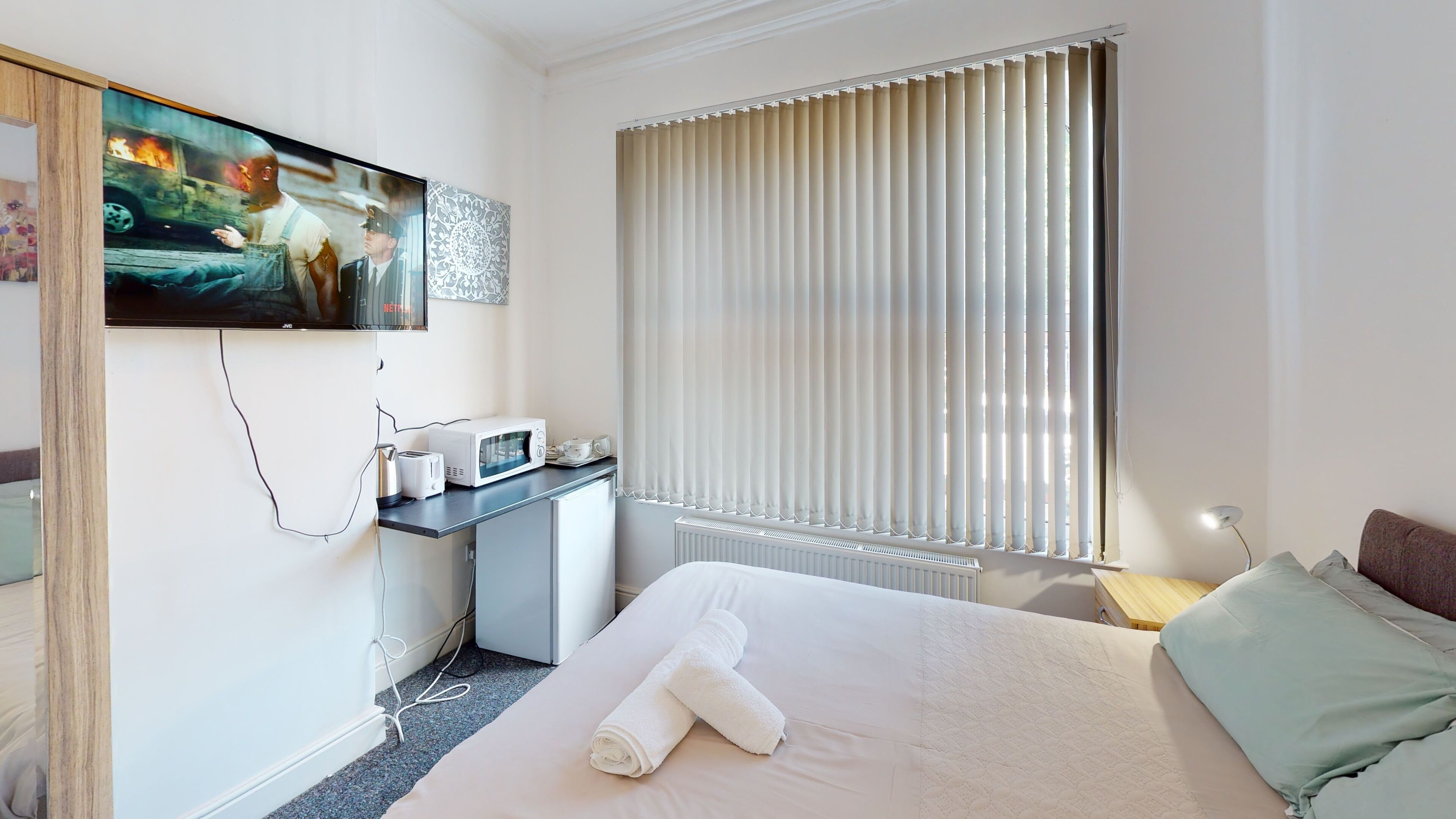 A hotel room with a bed, a desk with a microwave, a TV, and a window with blinds.