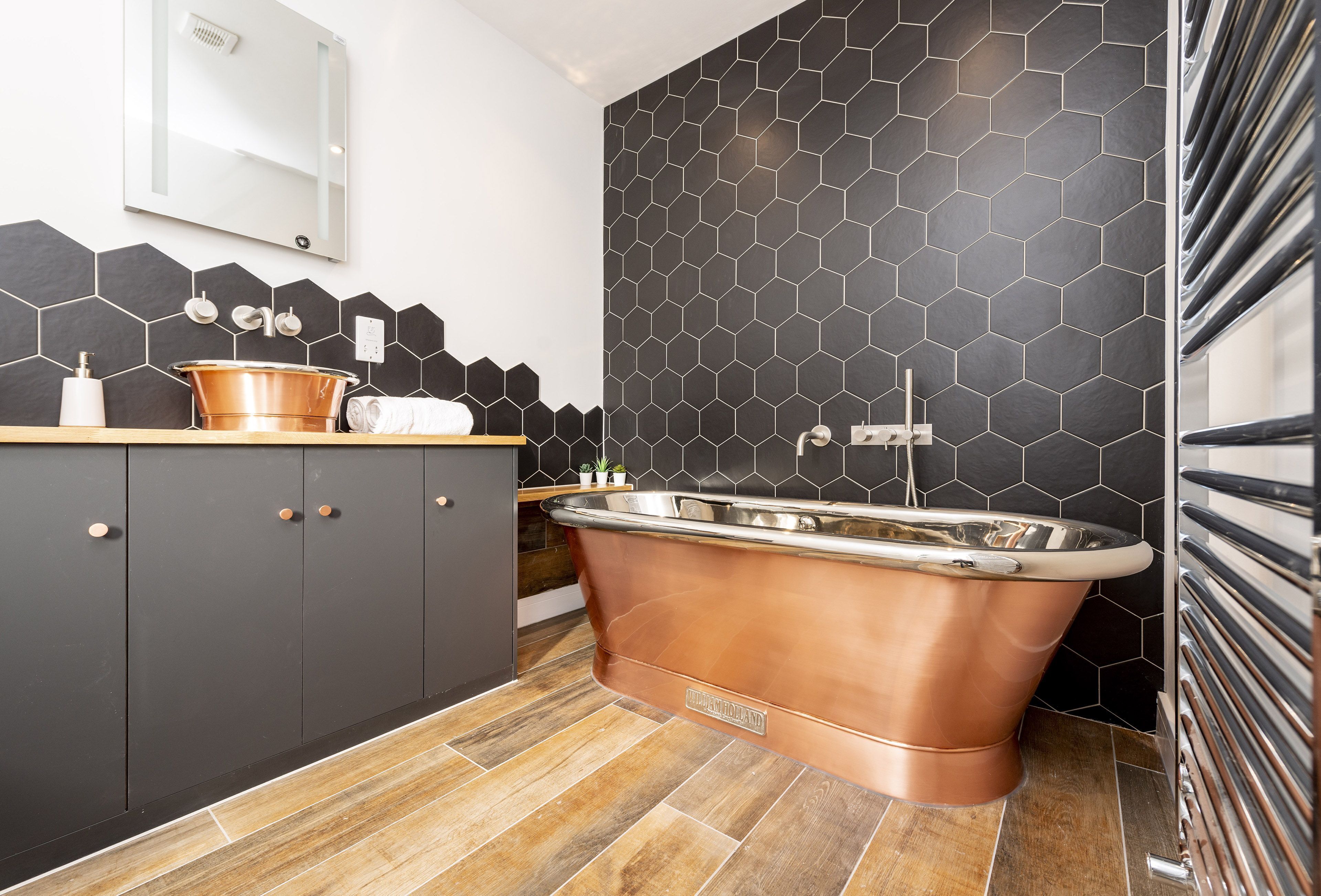 Une salle de bain moderne avec une baignoire en cuivre sur pieds, des carreaux hexagonaux noirs et un plancher en bois.