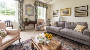 Un salón con sofá, sillones, una mesa de centro con tetera y flores, un espejo y cuadros en la pared.