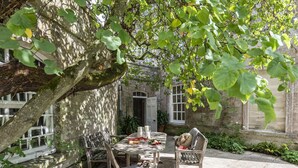 Un edificio de piedra con una mesa y sillas en el patio, rodeado de follaje y un árbol.