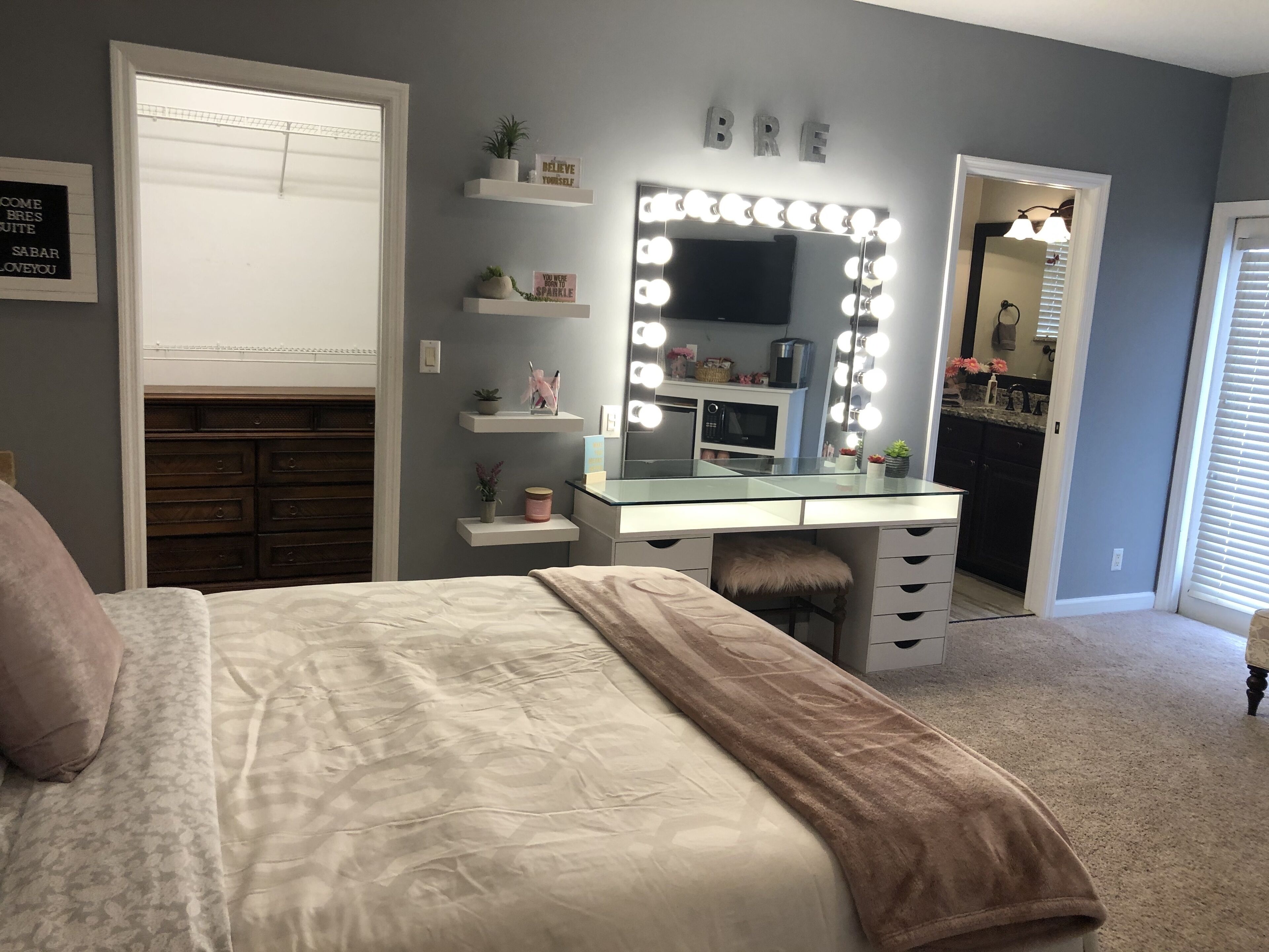 A bedroom with a bed, a vanity with a mirror and lights, a dresser, and shelves with decorative items.