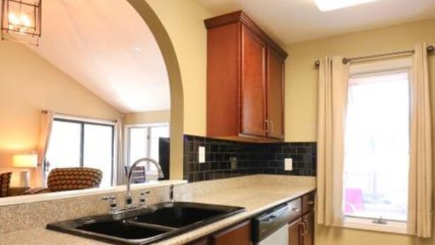 Una cocina con un lavamanos doble, armarios de madera, una ventana con cortinas y piso de baldosas.