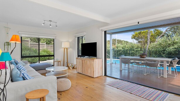 A modern living room with a large TV, a sofa, a coffee table, and a dining area with a pool view.