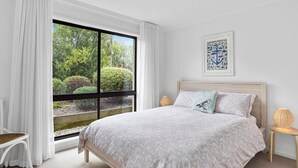 A bedroom with a large window, a bed with a patterned bedspread, a wall-mounted artwork, and a bedside lamp.