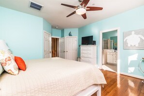 Une chambre à coucher avec un lit, un ventilateur de plafond, une télévision et une salle de bain visible à travers une porte ouverte.