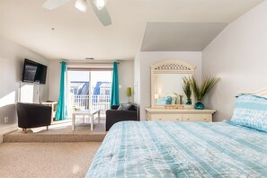 Ein Schlafzimmer mit Bett, Kommode, Fernseher, Stuhl und einem Balkon mit Vorhängen.