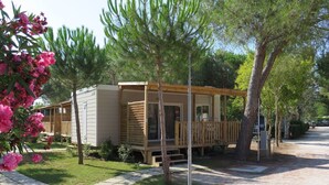 A small wooden cabin with a deck, surrounded by trees and a gravel path. - Lovely apartment in villa for 5 people with A/C, WIFI, pool, terrace and pets allowed (Bibbona)