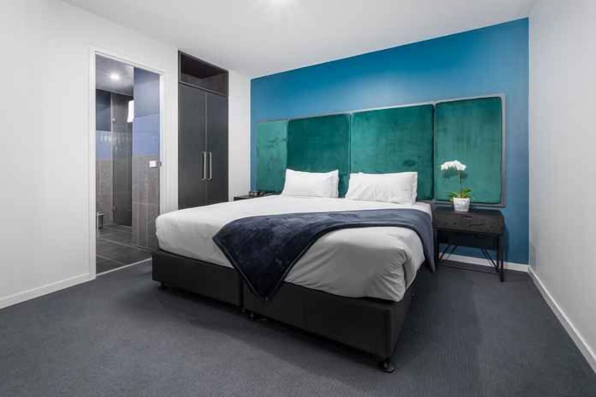 A modern bedroom with a large bed, a dark headboard, and a small bedside table with a plant.