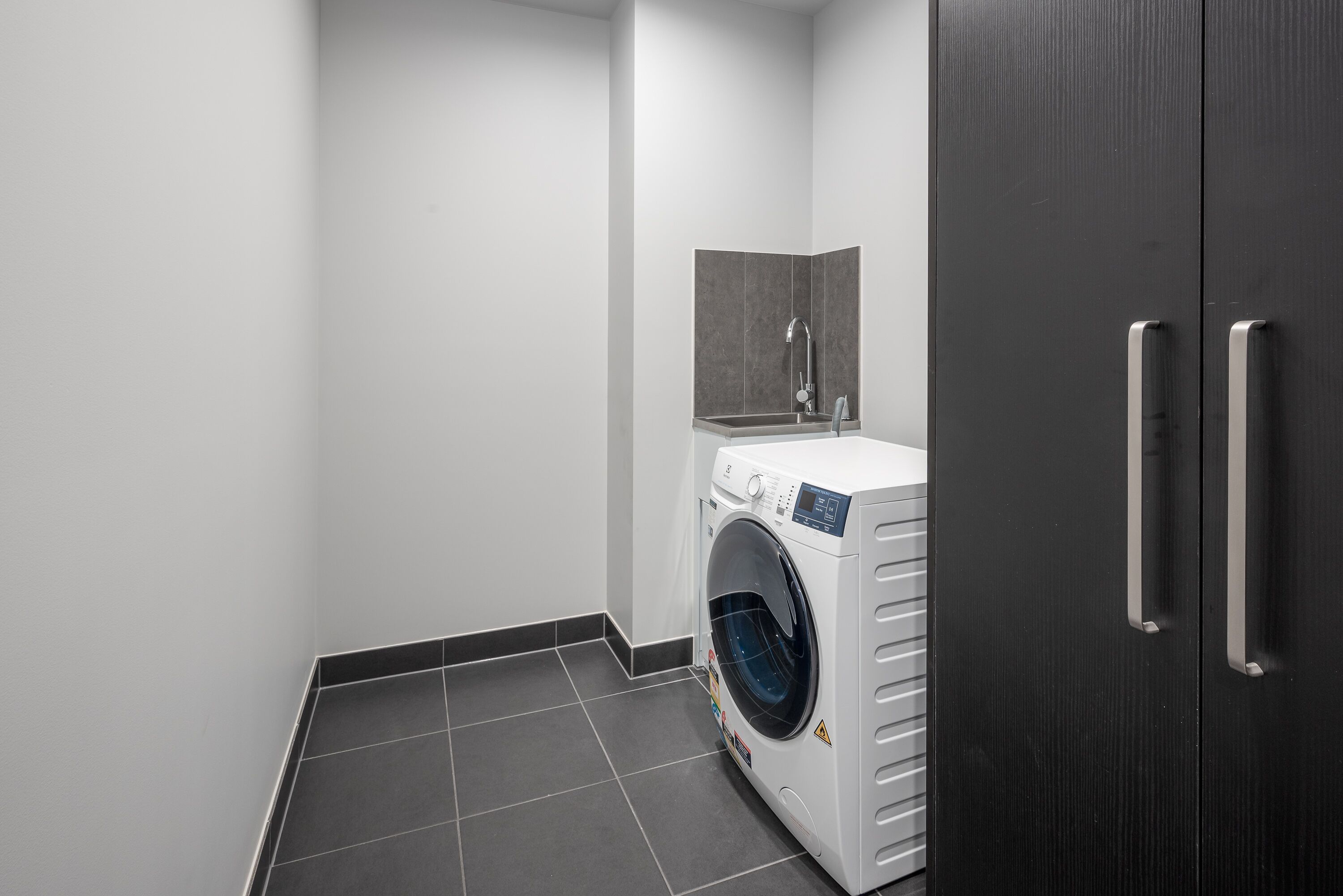 A compact laundry room with a front-loading washing machine, a small sink, and a dark wooden wardrobe.