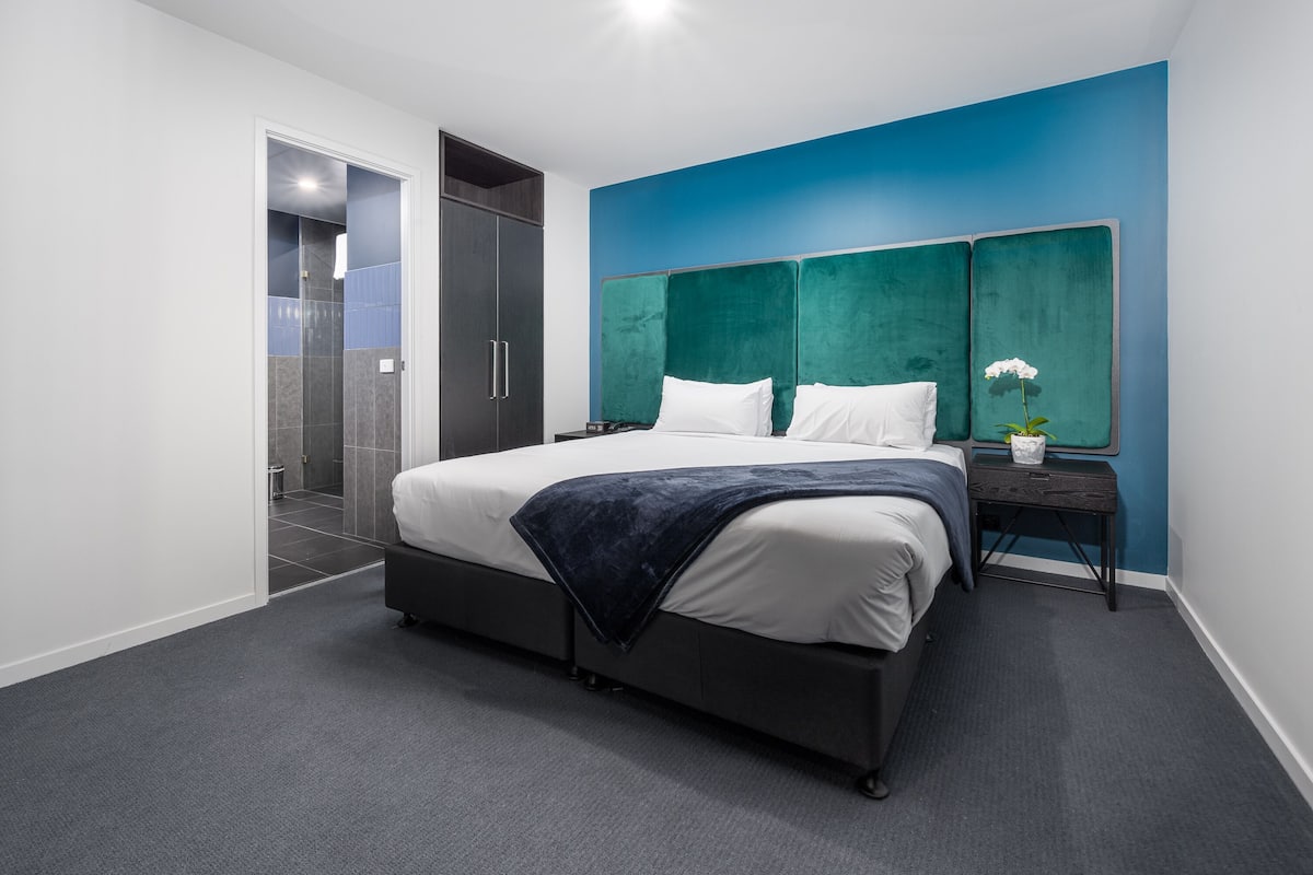 Una camera da letto moderna con un letto grande, un comodino, una TV a muro e un bagno visibile attraverso una porta aperta.
