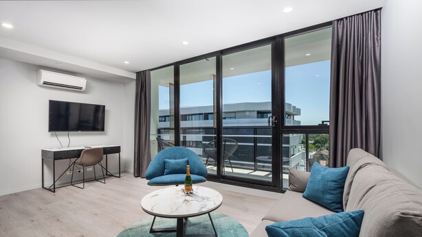 Ein modernes Wohnzimmer mit einer Couch, einem Couchtisch, einem Fernseher und einem Balkon mit Blick auf Gebäude.