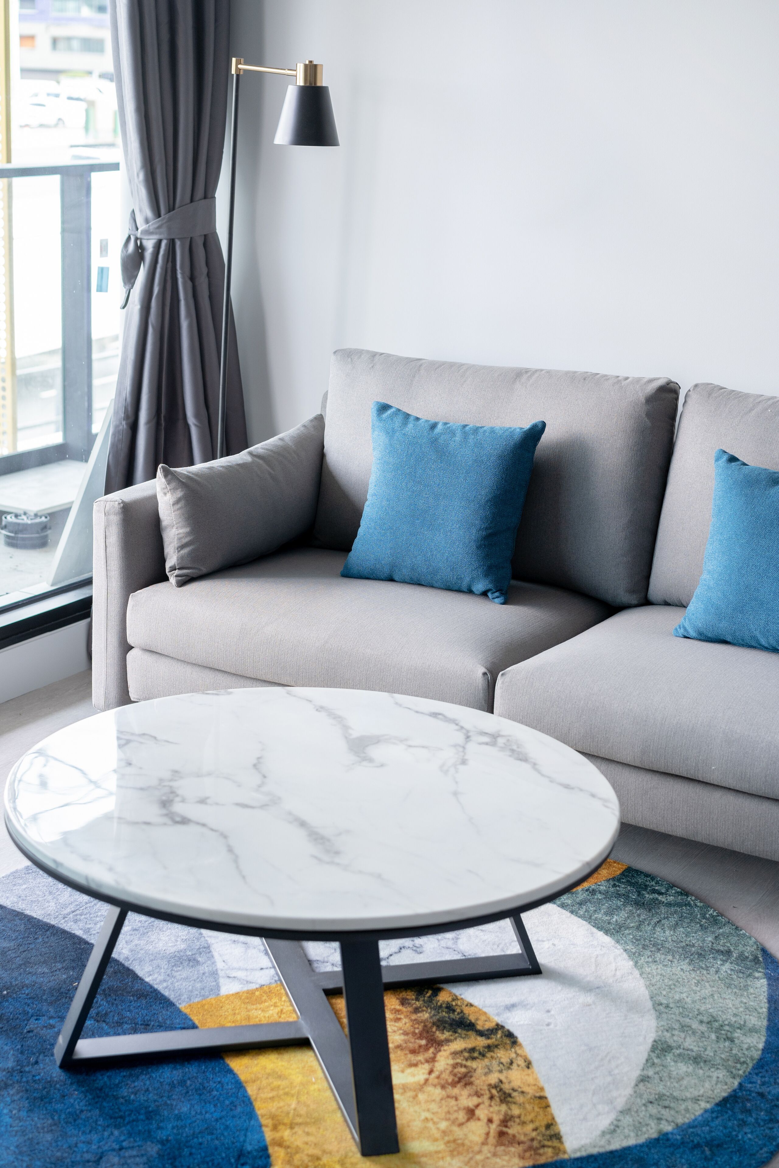 Un salon moderne avec un canapé gris, des coussins bleus, une table basse ronde en marbre et un tapis coloré.