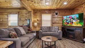A cozy log cabin interior with two grey sofas, a coffee table, a fireplace, and a large TV screen displaying animated characters. - Smoky Mountain Escape · Mini Golf · Private Hot Tub · Close to Dollywood (Sevierville)