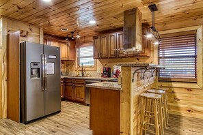Una cocina con armarios de madera, un refrigerador de acero inoxidable, una barra con taburetes y un ventanal con persianas.