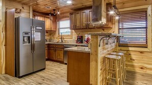 A wooden cabin-style kitchen with a stainless steel refrigerator, wooden cabinets, a built-in island with stools, and a large window with blinds. - Smoky Mountain Escape · Mini Golf · Private Hot Tub · Close to Dollywood (Sevierville)