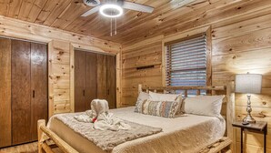 A wooden cabin bedroom with a bed, a ceiling fan, a window with blinds, and a bedside table with a lamp. - Smoky Mountain Escape · Mini Golf · Private Hot Tub · Close to Dollywood (Sevierville)