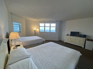 A hotel room with two beds, a desk, a dresser, a TV, and a window with a view of the sea.