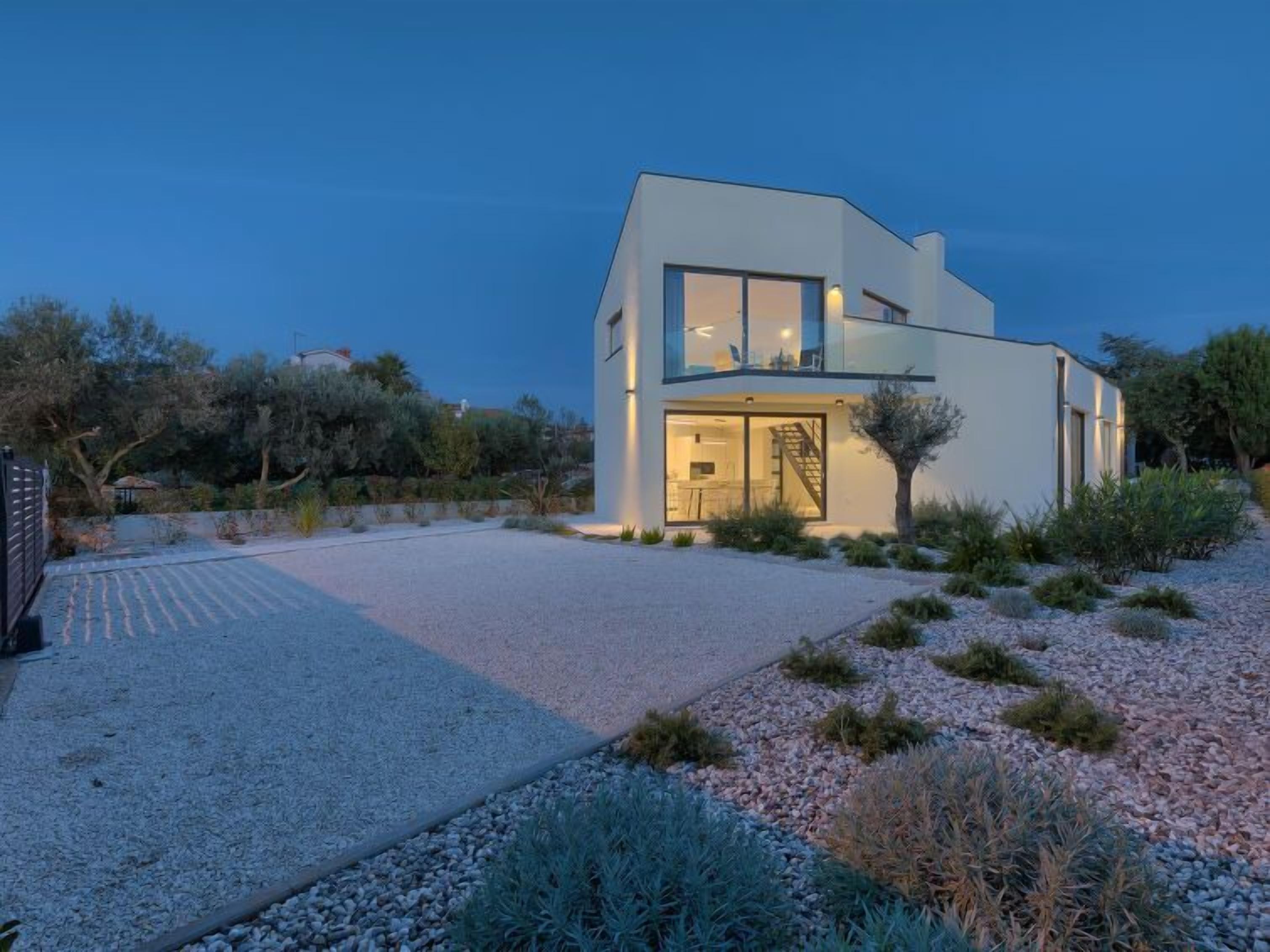 A modern two-story house with a large glass door entrance, a visible staircase, and a gravel driveway.