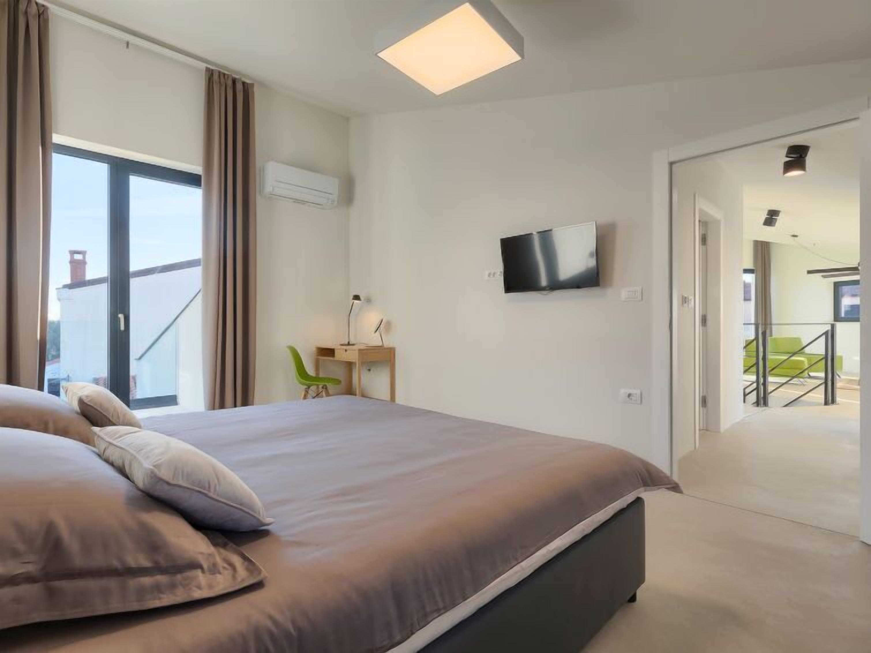 A modern hotel room with a large bed, a desk with a chair, a flat-screen TV, and a balcony with a view.