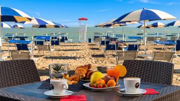 Ein Tisch am Strand mit Frühstücksgebäck, ein Glas Orangensaft und Blick auf den Strand mit Sonnenschirmen und Liegen.