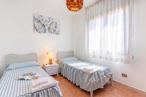 Una camera da letto con due letti singoli, un comodino con una lampada, una finestra con tende leggere e un'opera d'arte appesa al muro.