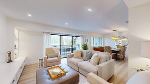 A modern living room with a sofa set, a dining table with chairs, and a large sliding glass door leading to an outdoor area.
