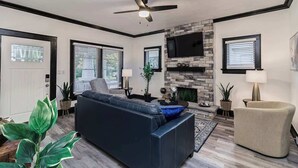 A modern living room with a dark blue sofa, a beige armchair, a fireplace, and a ceiling fan. - Historic Home Steps from Lake Hollingsworth, (Lakeland)