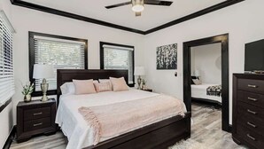 A bedroom with a large bed, a ceiling fan, two windows with blinds, a dresser, and a mirror. - Historic Home Steps from Lake Hollingsworth, (Lakeland)