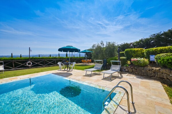 Una piscina con sillas de descanso y sombrillas, rodeada por un patio de piedra y vistas al paisaje.