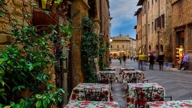 Una calle estrecha con mesas y sillas cubiertas con manteles florales, indicando un área de comedor al aire libre.
