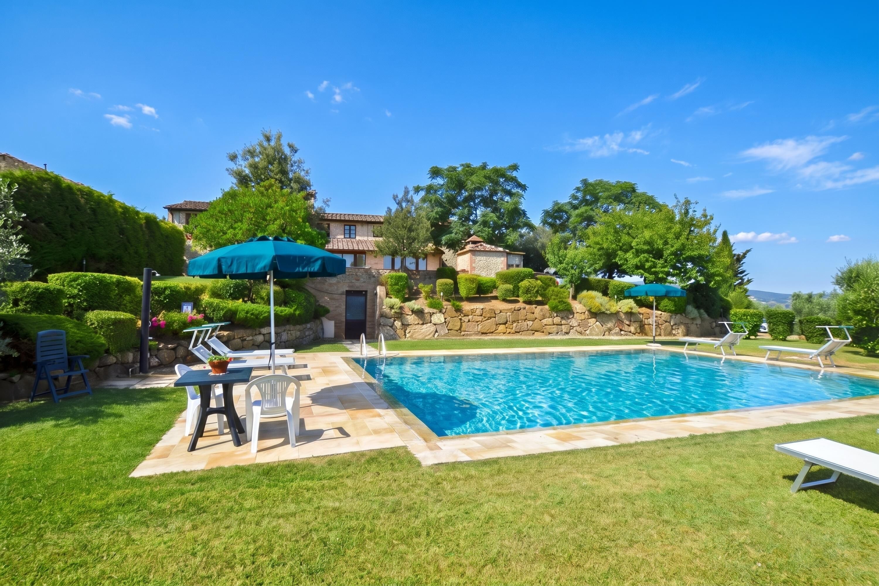 Une piscine entourée de chaises longues et de parasols, avec une pelouse bien entretenue et des murs en pierre.