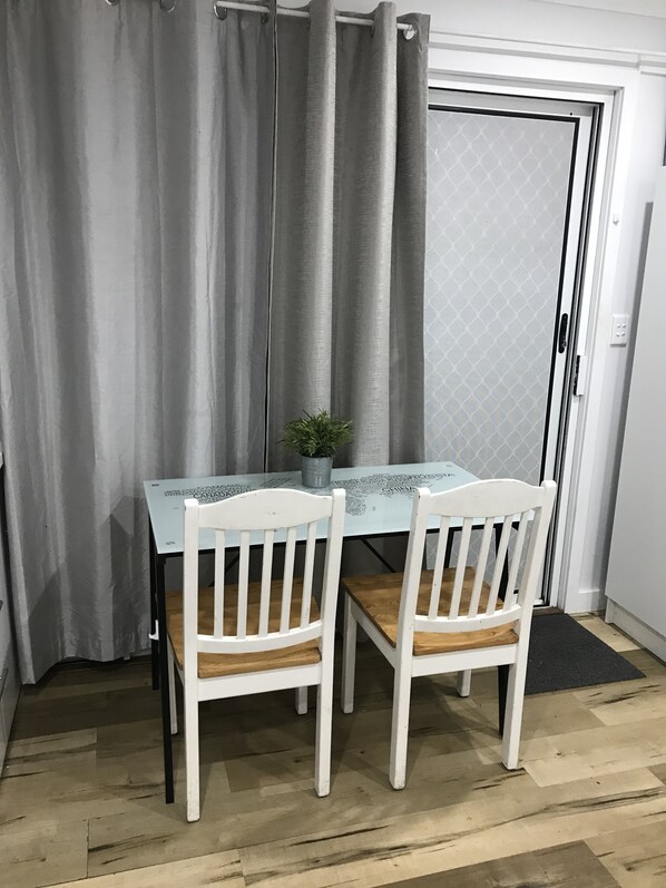 A dining area with a glass table, two white chairs, a potted plant, and a view through a door with a patterned screen.