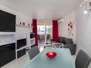Uma sala de estar moderna com um sofá cinzento, uma mesa de jantar de vidro com uma tigela vermelha e uma TV sobre um móvel preto.