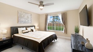 Een slaapkamer met een groot bed, een plafondventilator, een raam met uitzicht, een flatscreen-tv en een nachtkastje met een lamp.