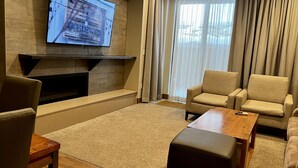 Una sala de estar moderna con un televisor de pantalla plana montado en una pared de piedra, una chimenea, sillones color beige, una mesa de centro de madera y un ventanal con cortinas ligeras.