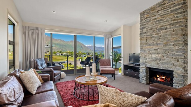 A spacious living room with a stone fireplace, leather sofas, and a large window overlooking a scenic landscape. - Kings View - Wanaka Holiday Home (Wanaka)