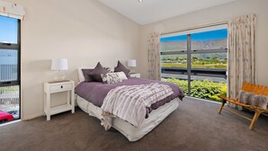 A bedroom with a large bed, two bedside tables, a chair, and a view of a landscape through a large window. - Kings View - Wanaka Holiday Home (Wanaka)