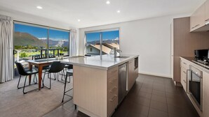 A modern kitchen with a large island, a dining area with chairs, and large windows offering a mountain view. - Kings View - Wanaka Holiday Home (Wanaka)