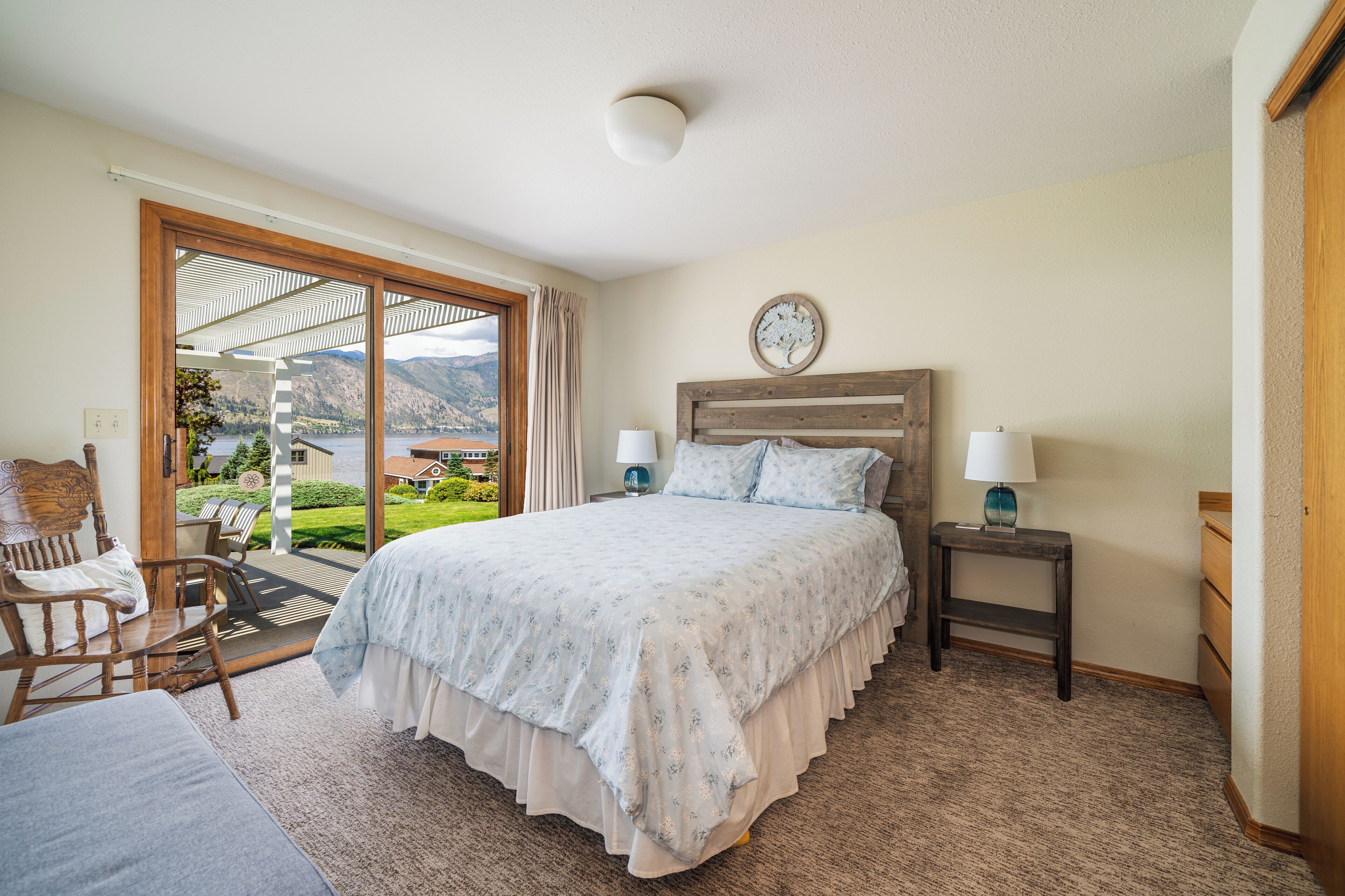 A bedroom with a large bed, two bedside tables with lamps, a wooden headboard, and a view of a lake through a sliding glass door.