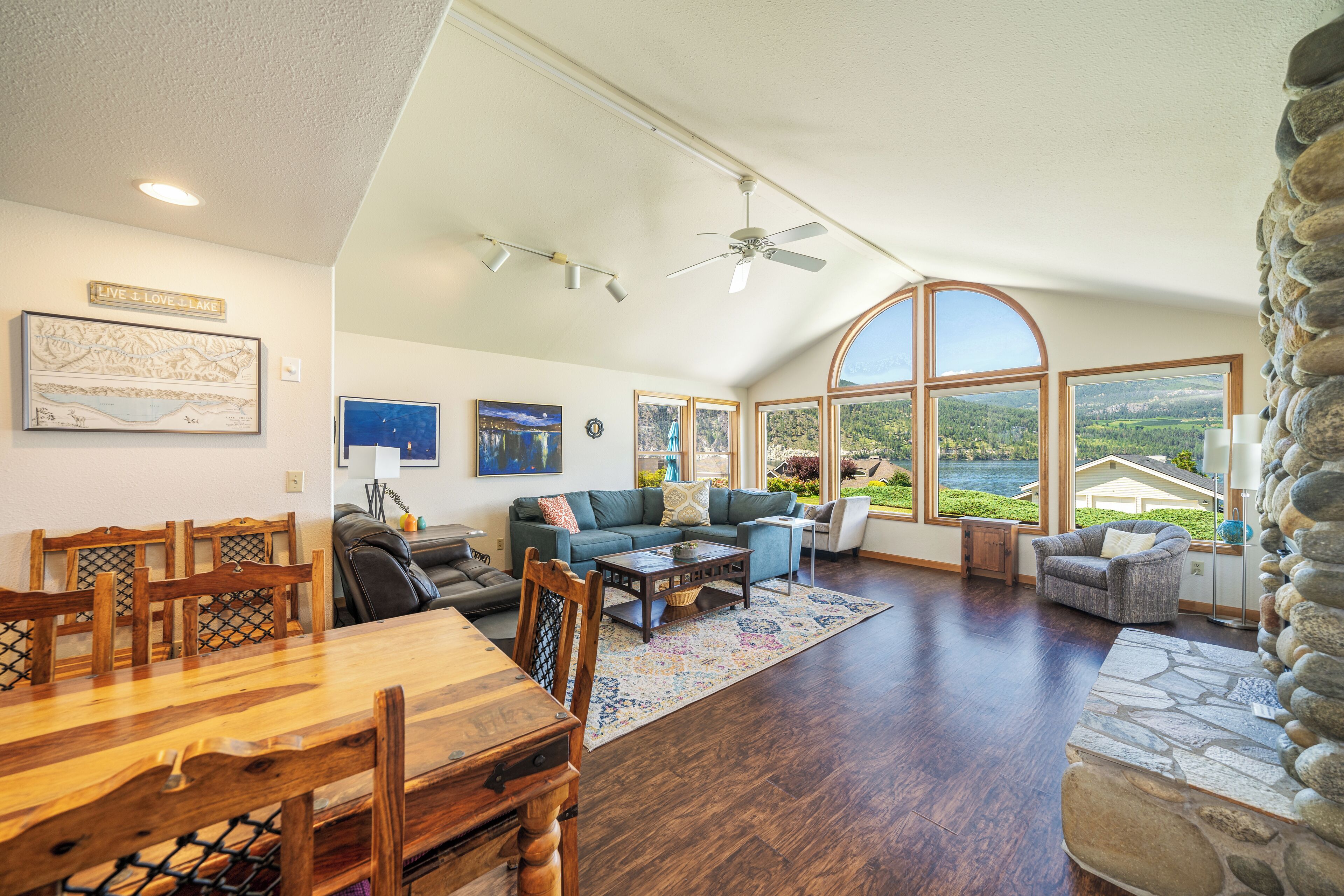 A spacious living room with a dining area, large windows offering a view of the outdoors, a ceiling fan, and a stone accent wall.
