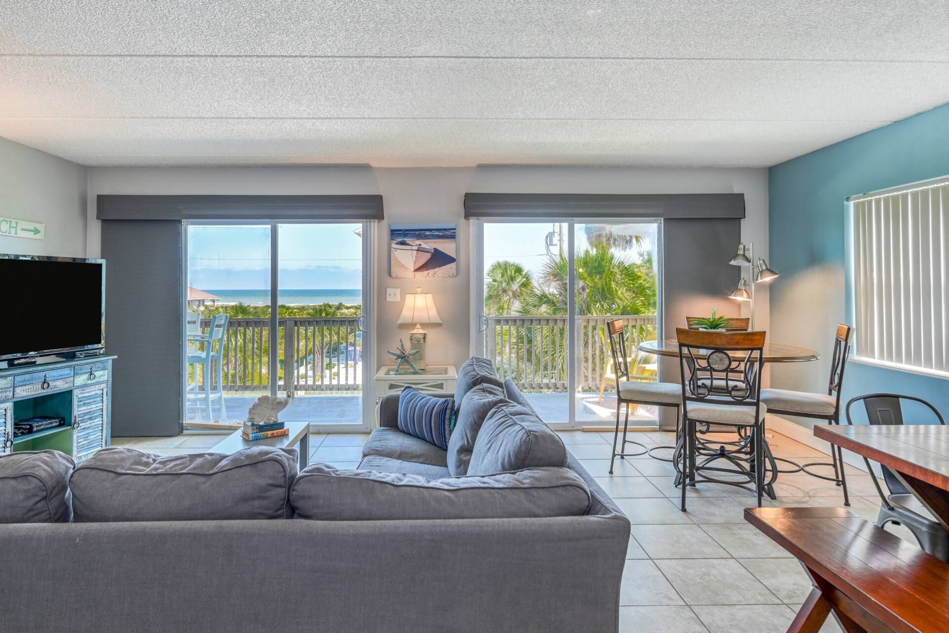 A living room with a sofa, a TV, a dining table with chairs, and a balcony with a view of the beach.