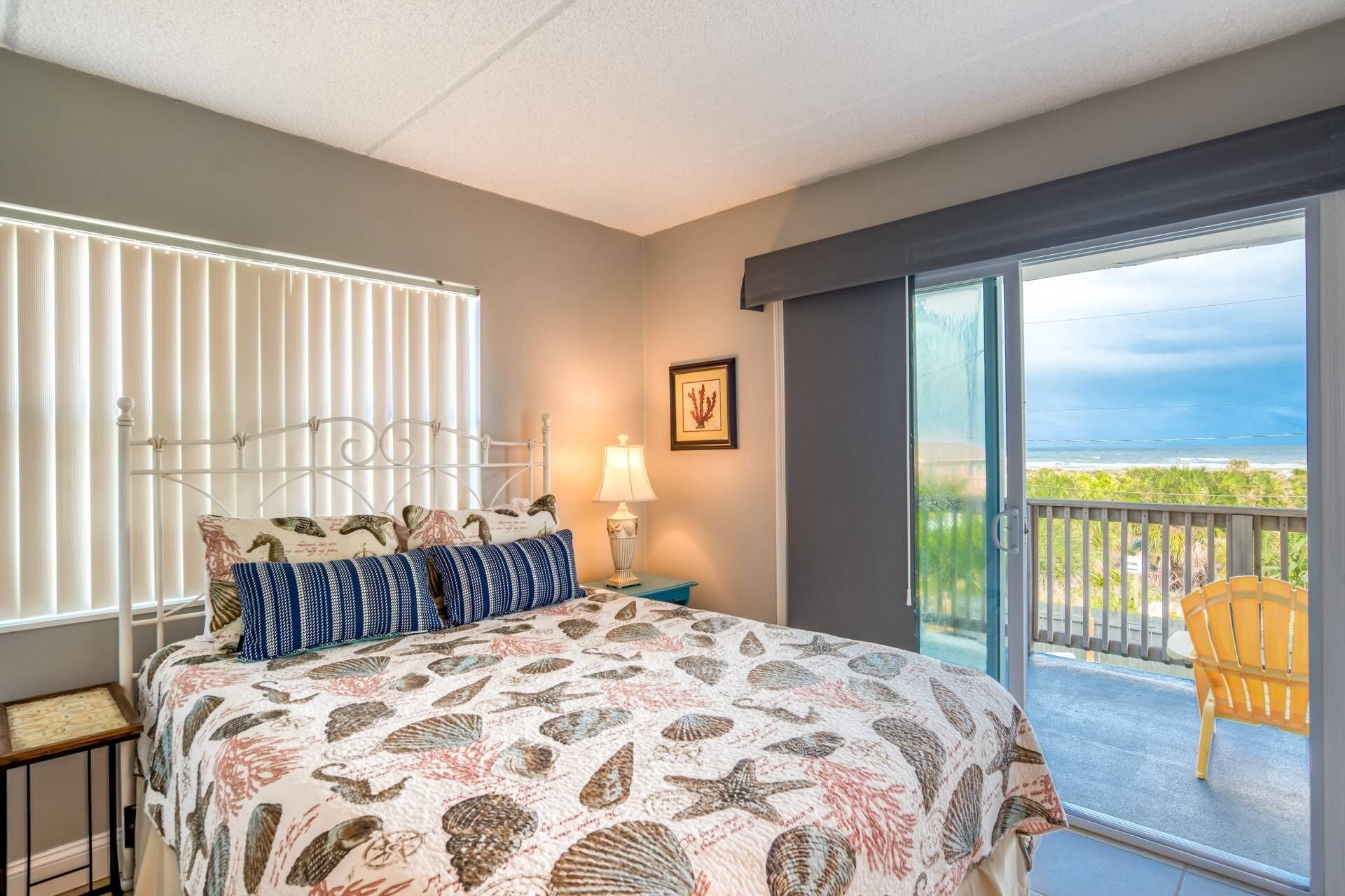 Une chambre à coucher comprenant un lit, une table de chevet, une lampe, un tableau encadré et une vue sur l’océan, visible à travers une porte-fenêtre coulissante.