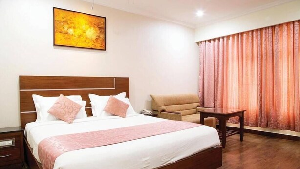 A hotel room with a large bed, a chair, a small table, and a window with curtains. - Ramyas Hotels (Tiruchirappalli)