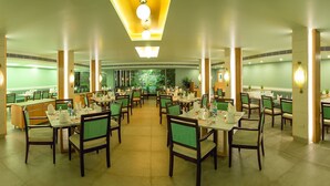 A spacious dining area with multiple tables and chairs, a central decorative wall, and a ceiling with recessed lighting. - Ramyas Hotels (Tiruchirappalli)